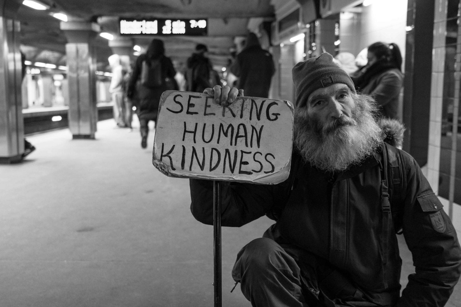 Photograph of a homeless man with a sign that says seeking human kindness to illustrate ways we can help the homeless