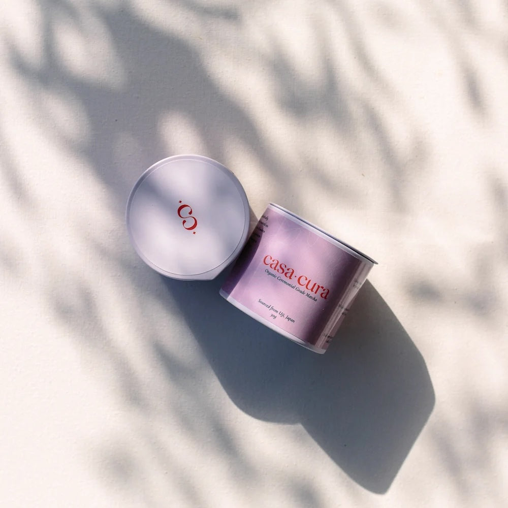 A jar and its lid from the brand Casa Cura, styled on a light beige surface with soft leaf shadows. The jar is lavender purple with red lettering, and the lid is a matching light purple with a red logo.