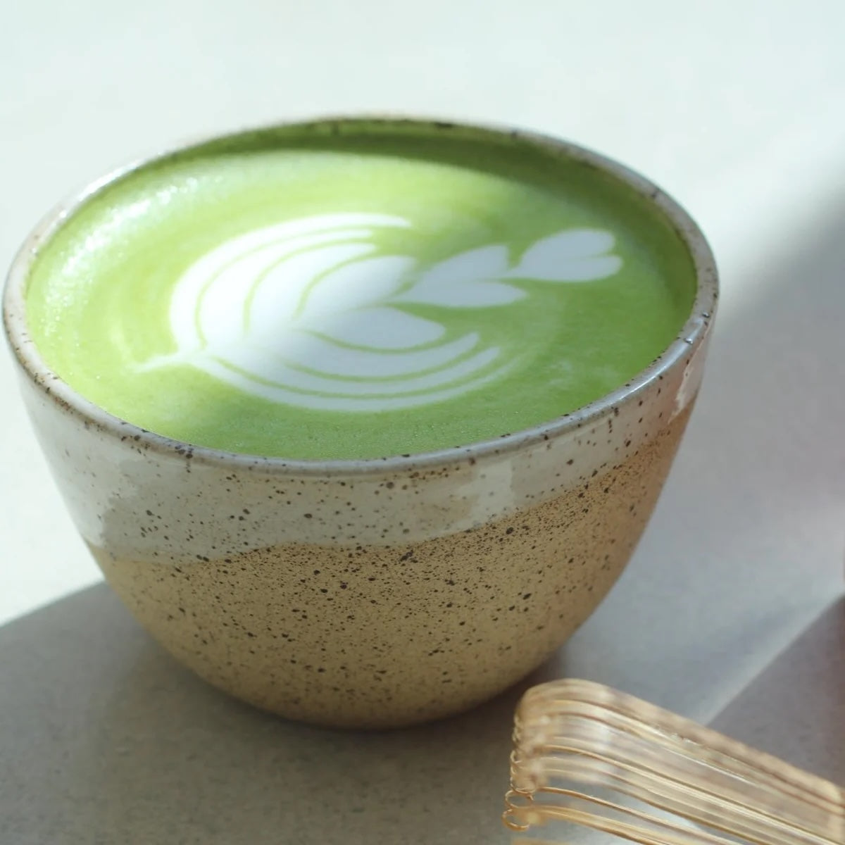 A close-up of a ceramic cup filled with green matcha latte art, set on a neutral surface with a small gold-toned accessory in the foreground.
