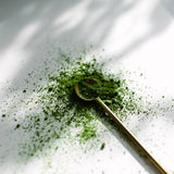 A small gold spoon filled with and surrounded by bright green powder scattered on a white surface, with soft leaf shadows cast across the scene.