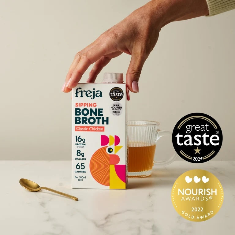 A hand holds a carton of Freja Sipping Bone Broth Classic Chicken next to a glass of broth and a gold spoon on a light marble surface. Two award badges are shown on the right: a black 'great taste 2024' badge and a gold 'Nourish Awards 2022 Gold Award' badge.