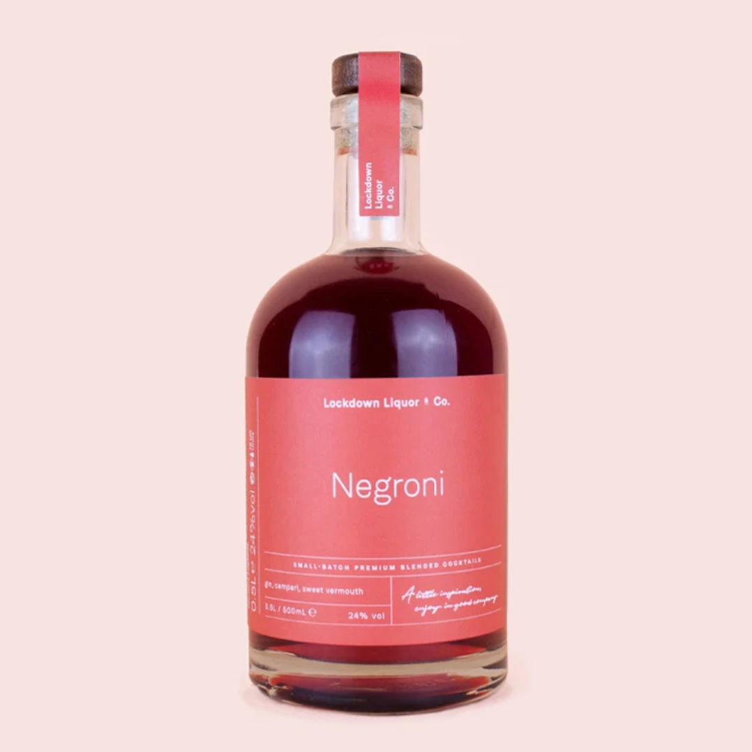 A bottle of Negroni liqueur by Lockdown Liquor & Co. The clear glass bottle is filled with a deep red-brown liquid and features a prominent dusty pink label with white text, sealed with a matching pink neck band and a dark cap, set against a solid pale pink background.