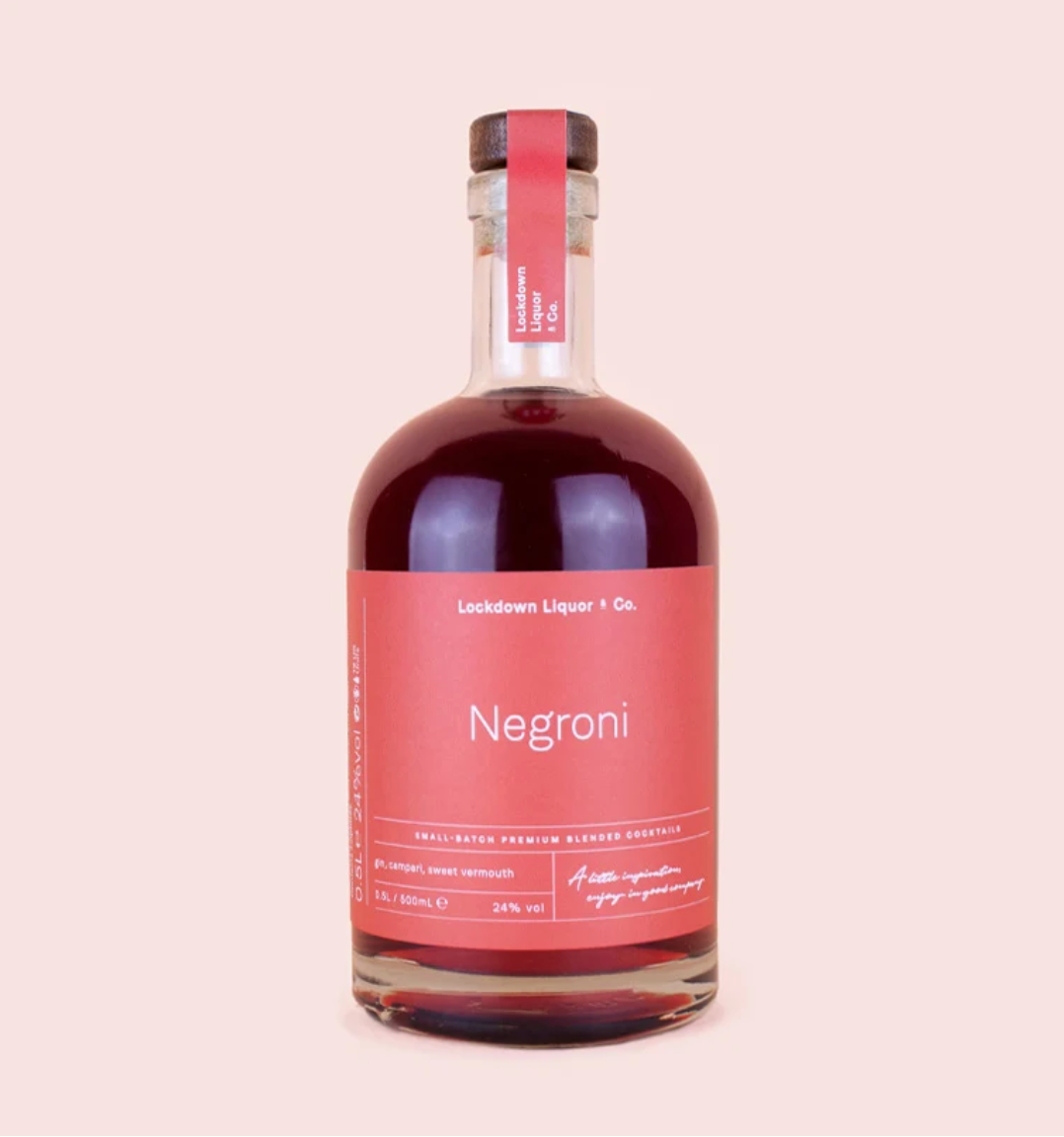 A bottle of Negroni liqueur by Lockdown Liquor & Co. The clear glass bottle is filled with a deep red-brown liquid and features a prominent dusty pink label with white text, sealed with a matching pink neck band and a dark cap, set against a solid pale pink background.