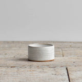 A small cylindrical ceramic container with a textured, speckled off-white glaze and a natural brown clay base, set on a rustic wooden surface against a neutral gray background.