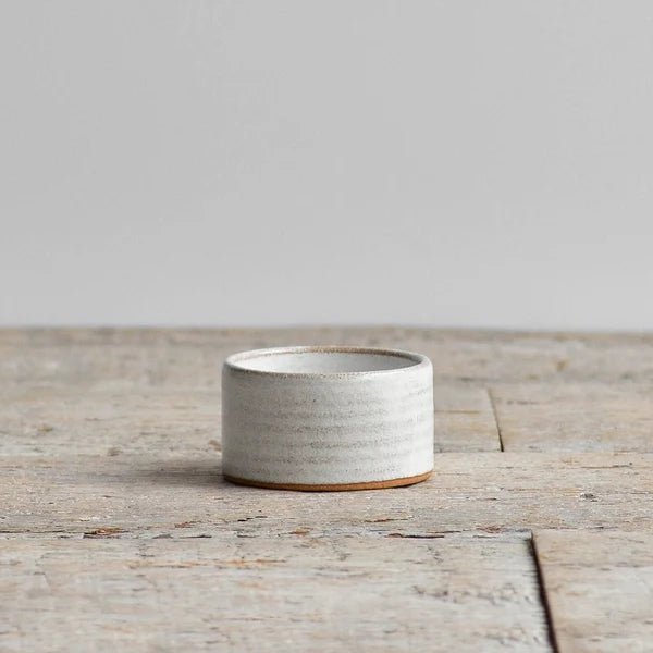 A small cylindrical ceramic container with a textured, speckled off-white glaze and a natural brown clay base, set on a rustic wooden surface against a neutral gray background.
