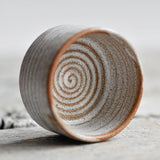 A handmade ceramic cup with a textured, speckled finish, featuring a spiral pattern inside and a terracotta-colored rim, resting on a rustic wooden surface.