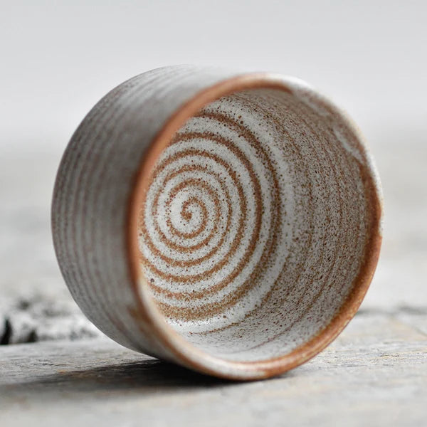 A handmade ceramic cup with a textured, speckled finish, featuring a spiral pattern inside and a terracotta-colored rim, resting on a rustic wooden surface.
