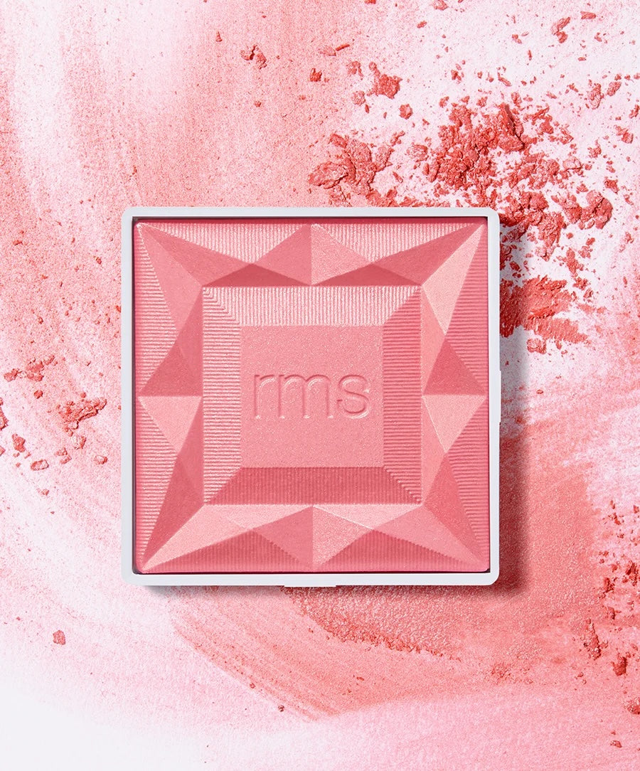 A square pink pressed powder makeup compact with a white border and the embossed brand name 'rms' in the center, placed on a textured pink background with loose powder scattered around.