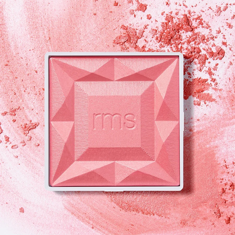 A square pink pressed powder makeup compact with a white border and the embossed brand name 'rms' in the center, placed on a textured pink background with loose powder scattered around.