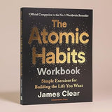 The Atomic Habits Workbook book by James Clear, featuring a black cover with gold and white text, photographed on a neutral beige background.