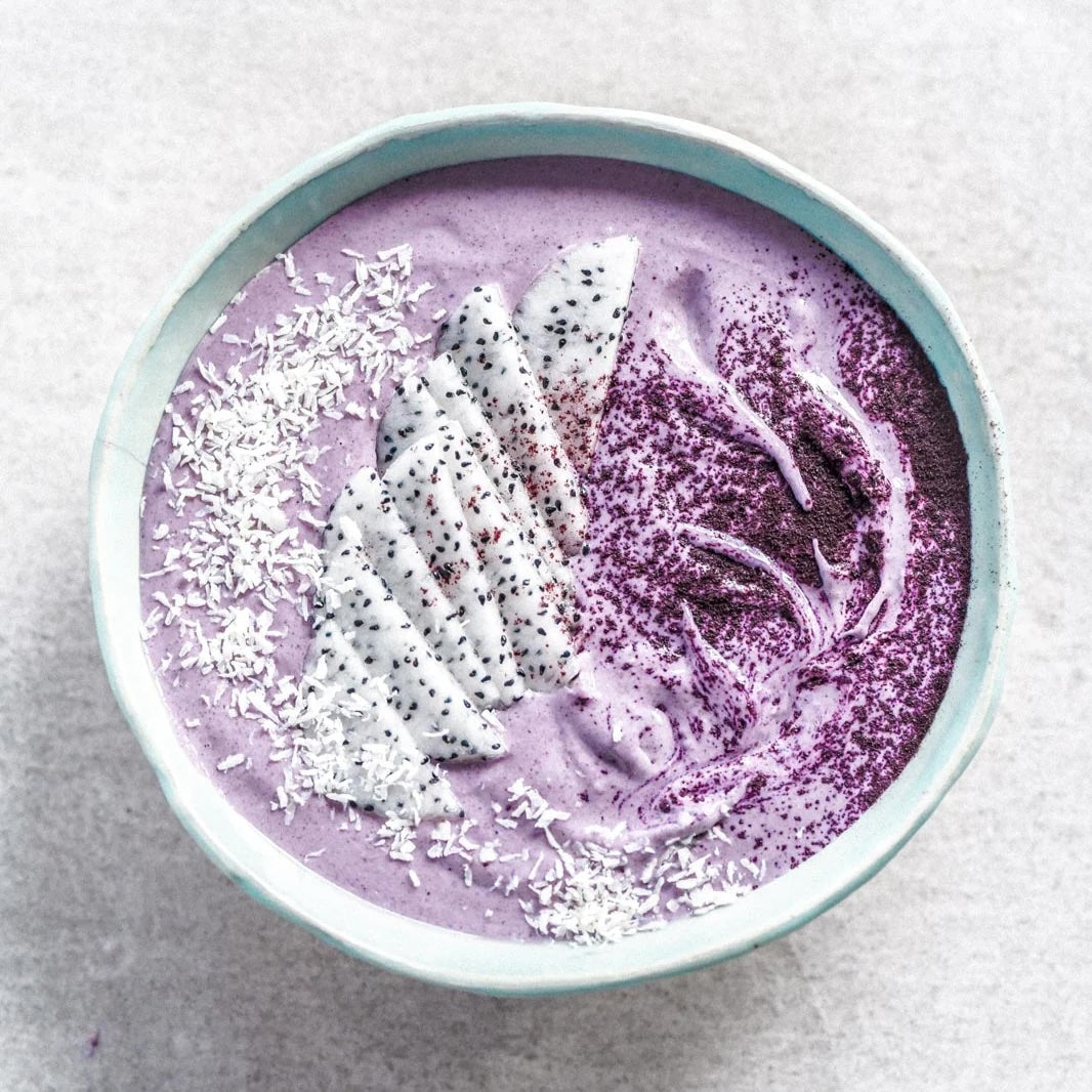 A top-down view of a light blue ceramic bowl filled with a purple smoothie or dessert, topped with white coconut flakes, a slice of white fruit with black seeds, and dark berry powder, set on a light gray textured surface.