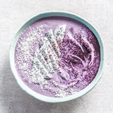 A top-down view of a light blue ceramic bowl filled with a purple smoothie or dessert, topped with white coconut flakes, a slice of white fruit with black seeds, and dark berry powder, set on a light gray textured surface.