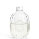 A clear ribbed glass bottle with a matching glass stopper, shown on a white background. The bottle has a circular embossed logo in the centre.