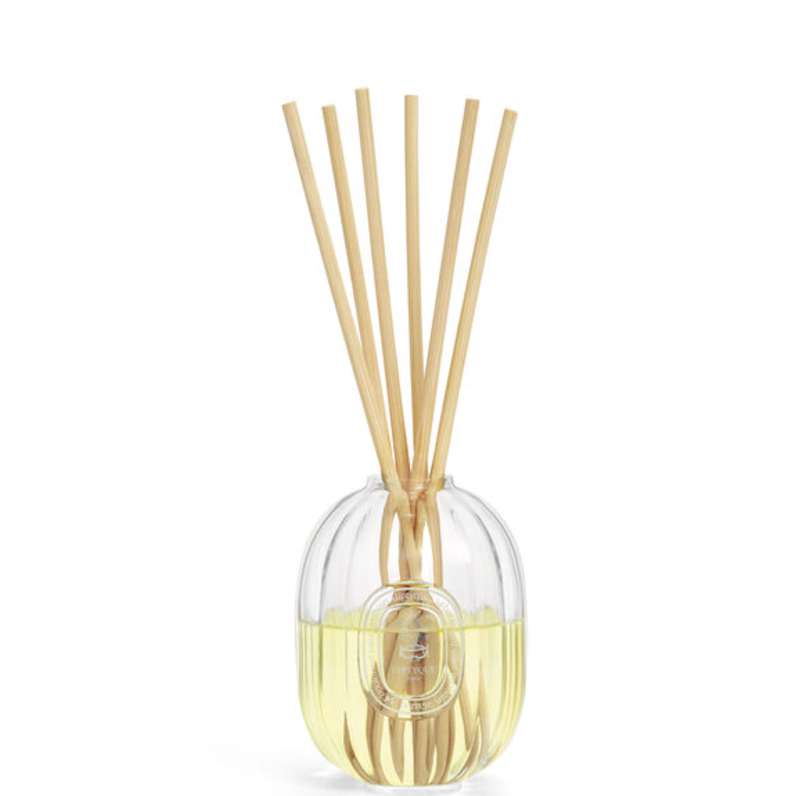 A reed diffuser with a clear ribbed glass bottle filled with pale yellow liquid, topped with six natural bamboo reeds, on a solid white background.