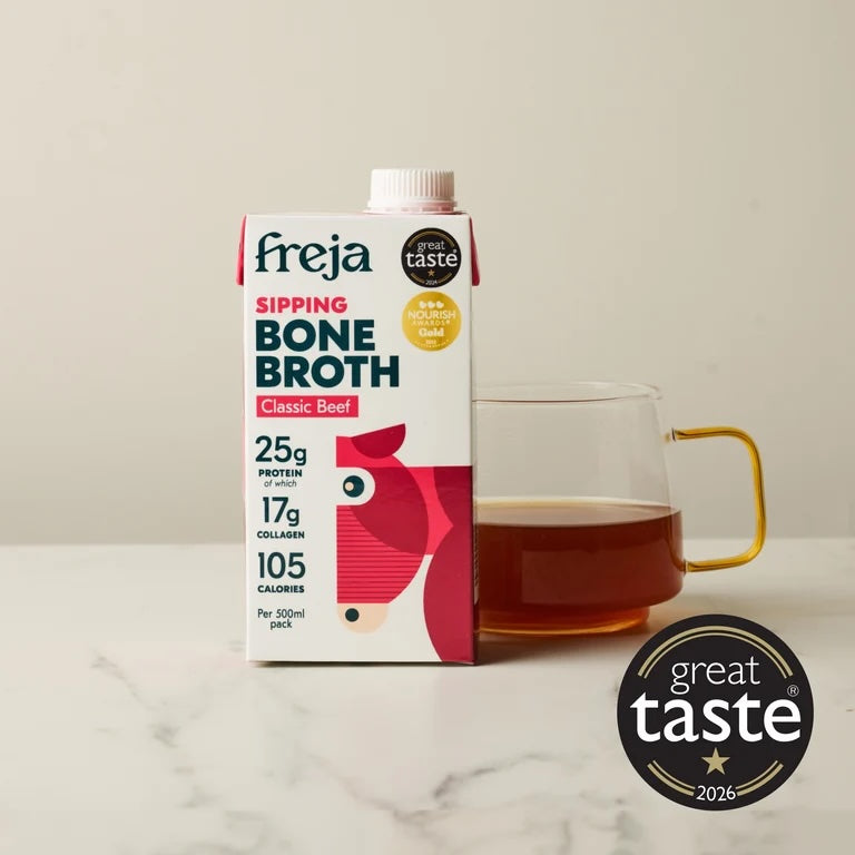 A carton of Freja Sipping Bone Broth Classic Beef stands on a light marble surface beside a clear glass mug filled with amber broth. The package is white with red and dark blue text and a stylized cow illustration.