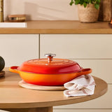 An orange and red enameled cast iron Dutch oven with a lid, set on a round wooden table in a modern kitchen setting with autumn vegetables and a white towel.