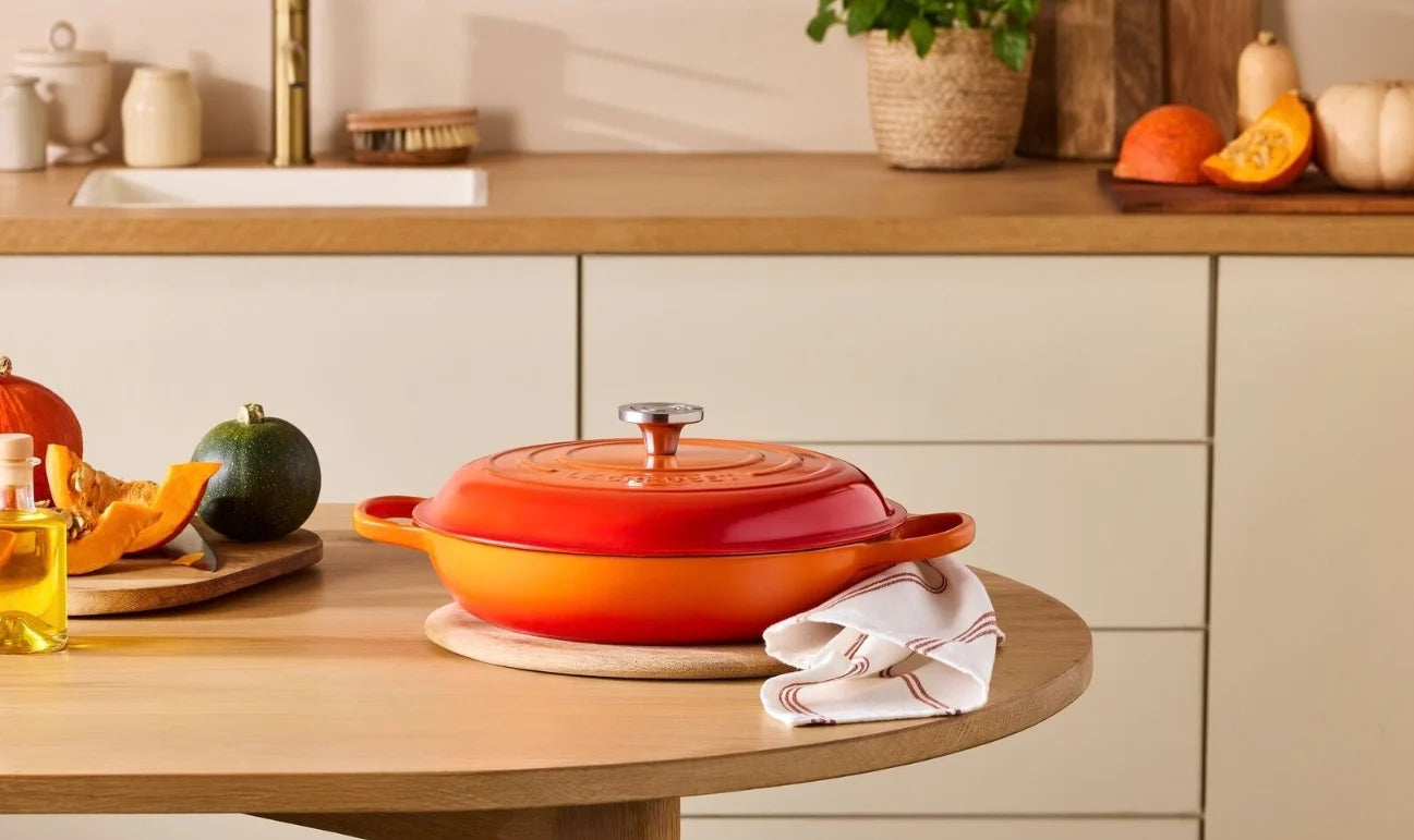 An orange and red enameled cast iron Dutch oven with a lid, set on a round wooden table in a modern kitchen setting with autumn vegetables and a white towel.
