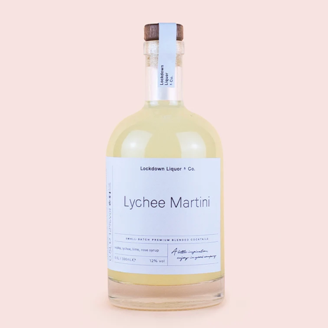 A clear glass bottle of Lockdown Liquor & Co. Lychee Martini with a white label and a dark brown wooden cap, set against a solid pale pink background.