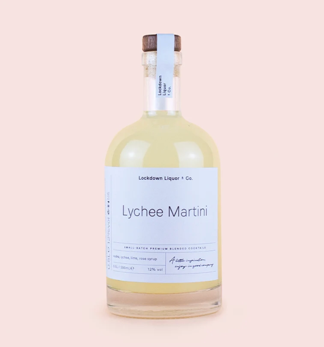A clear glass bottle of Lockdown Liquor & Co. Lychee Martini with a white label and a dark brown wooden cap, set against a solid pale pink background.
