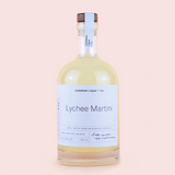 A clear glass bottle of Lockdown Liquor & Co. Lychee Martini with a white label and a dark brown wooden cap, set against a solid pale pink background.