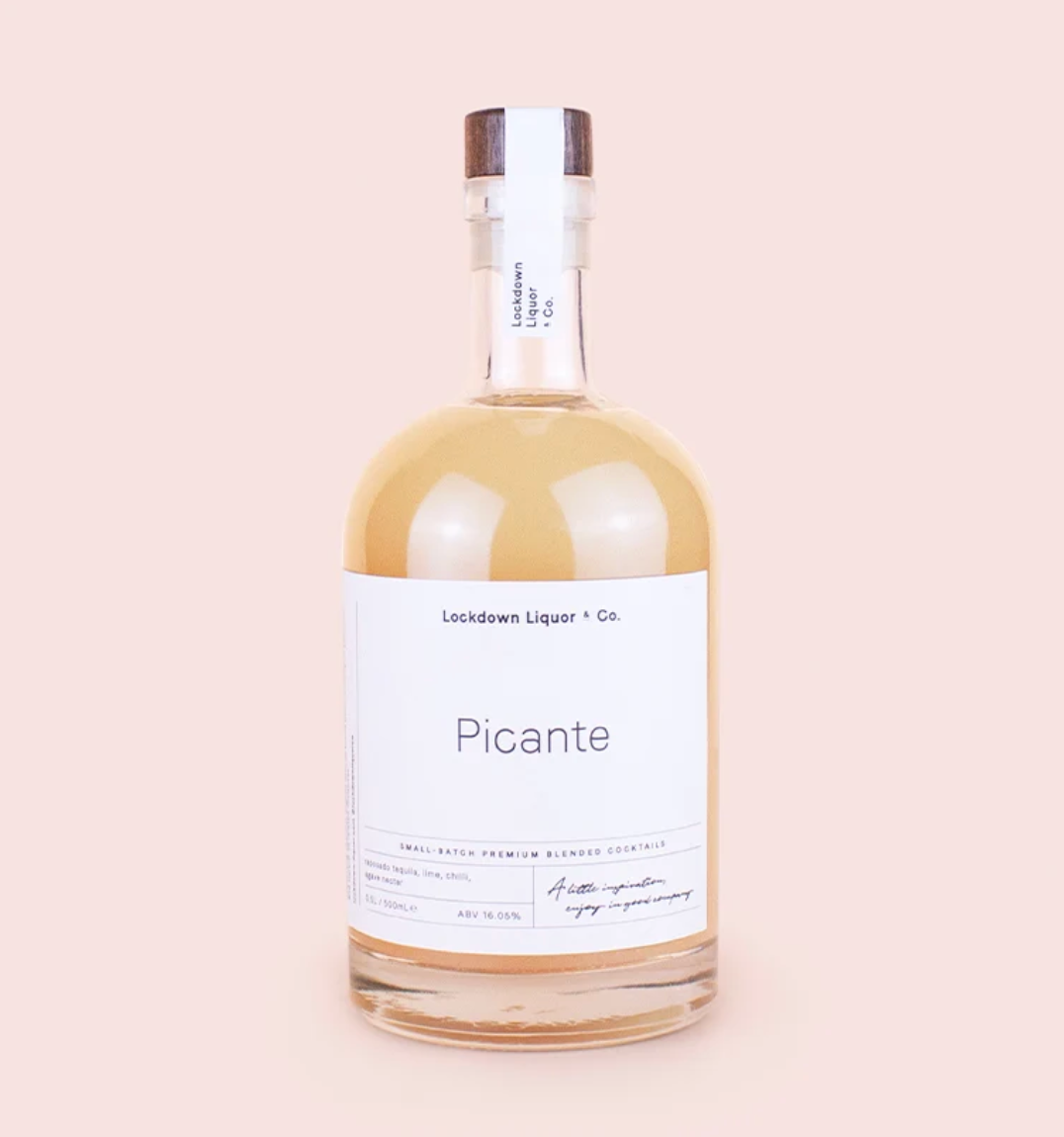 A bottle of Lockdown Liquor + Co. 'Picante' small batch premium blended cocktail mix, filled with a golden-amber liquid, featuring a white label and a dark brown wooden stopper, set against a solid pale pink background.