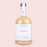 A bottle of Lockdown Liquor + Co. 'Picante' small batch premium blended cocktail mix, filled with a golden-amber liquid, featuring a white label and a dark brown wooden stopper, set against a solid pale pink background.