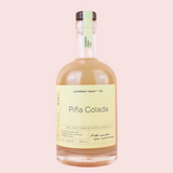 A bottle of Piña Colada by Lockdown Liquor & Co. featuring a pale yellow label and a light green neck seal, filled with a creamy beige liquid, set against a solid pale pink background.