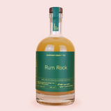 A bottle of Rum Rock by Lockdown Liquor & Co. featuring a dark green label and a brown cork stopper, filled with amber-coloured rum, set against a solid pale pink background.