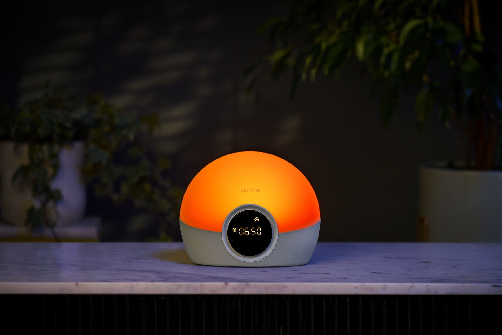 A dome-shaped smart alarm clock or lamp with a glowing orange top half and a light gray bottom half, sitting on a marble countertop in a dimly lit room with plants in the background.