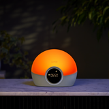 A dome-shaped smart alarm clock or lamp with a glowing orange top half and a light gray bottom half, sitting on a marble countertop in a dimly lit room with plants in the background.