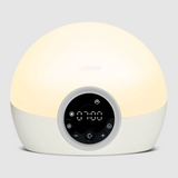 A dome-shaped Lumiie alarm clock lamp with a warm white glowing top half and a white bottom half, featuring a circular black digital control panel on the front.