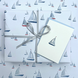 A gift-wrapped box and a matching greeting card with a sailboat print, tied with a gray ribbon, placed on top of matching patterned wrapping paper.