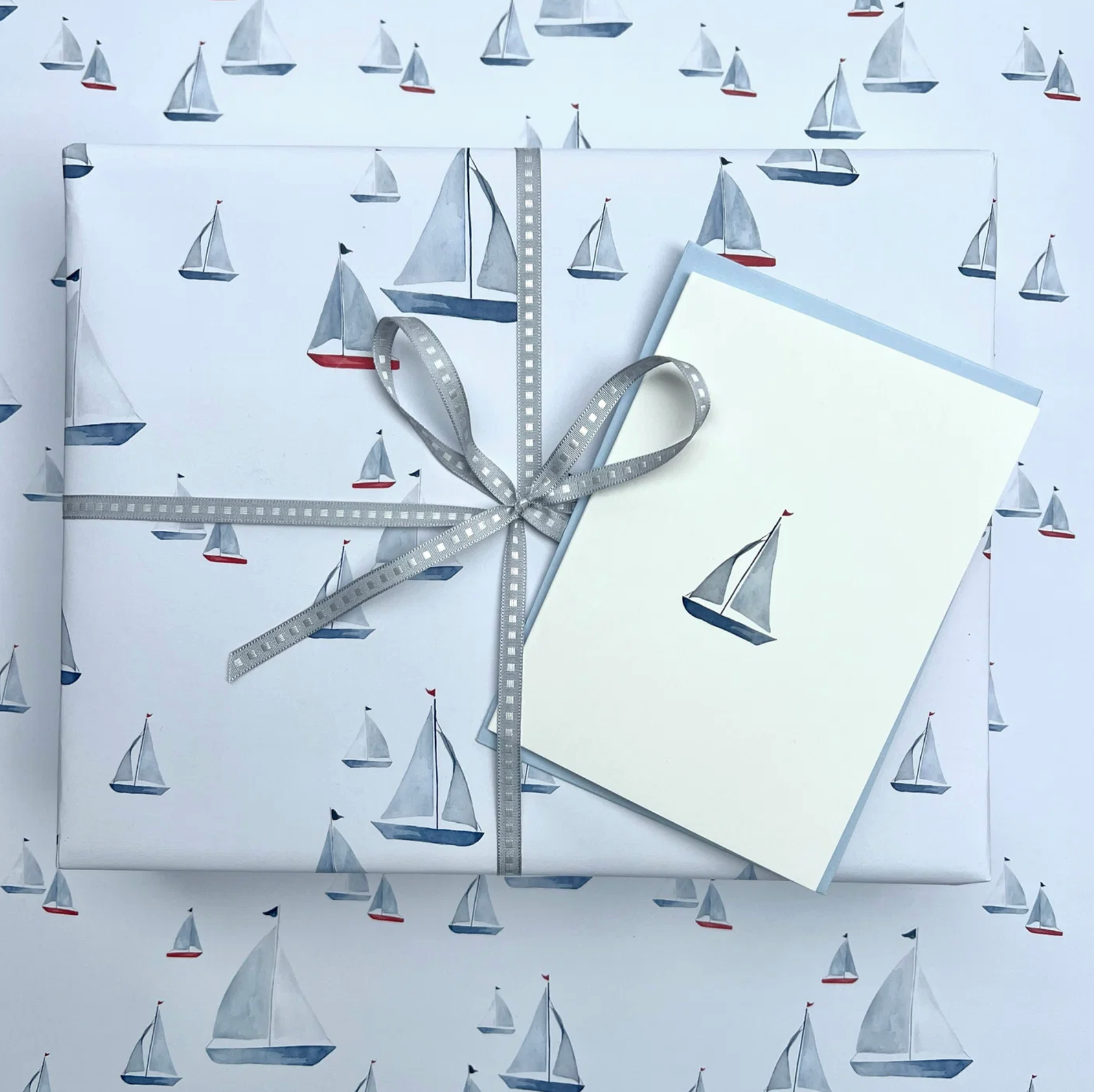 A gift-wrapped box and a matching greeting card with a sailboat print, tied with a gray ribbon, placed on top of matching patterned wrapping paper.