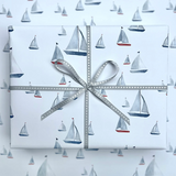 A square gift box wrapped in white paper printed with a repeating sailboat pattern, tied with a gray ribbon and bow in the center.