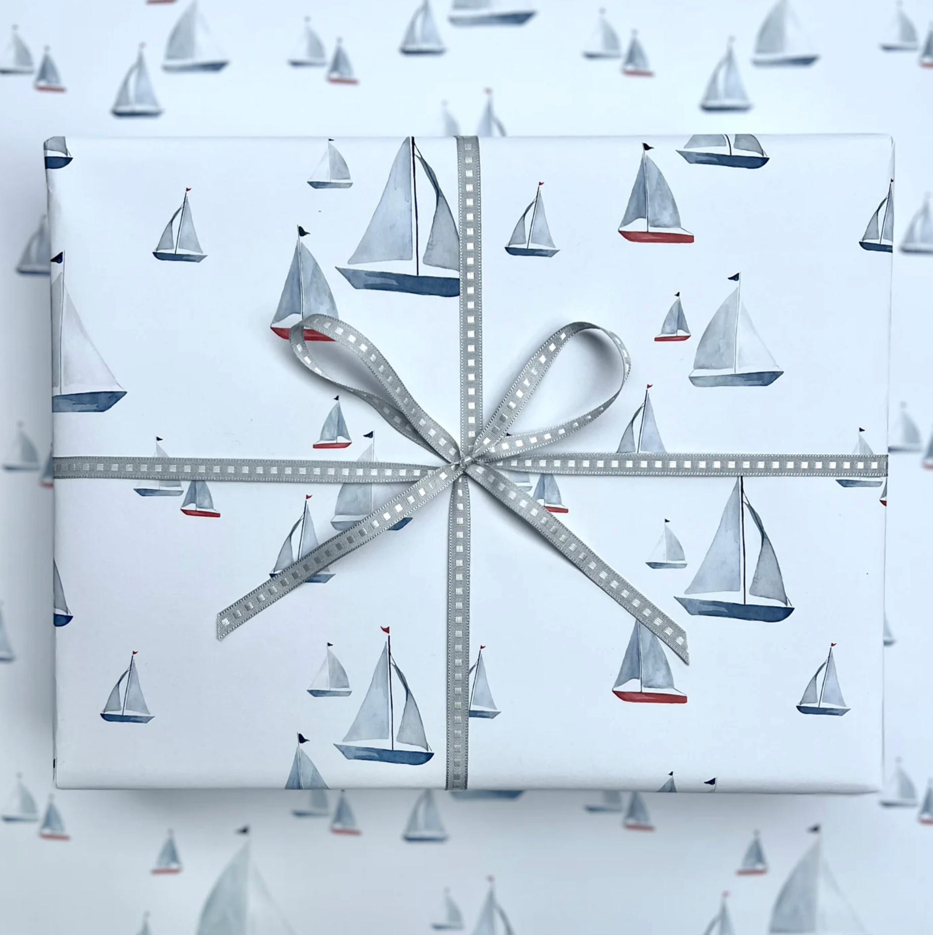 A square gift box wrapped in white paper printed with a repeating sailboat pattern, tied with a gray ribbon and bow in the center.