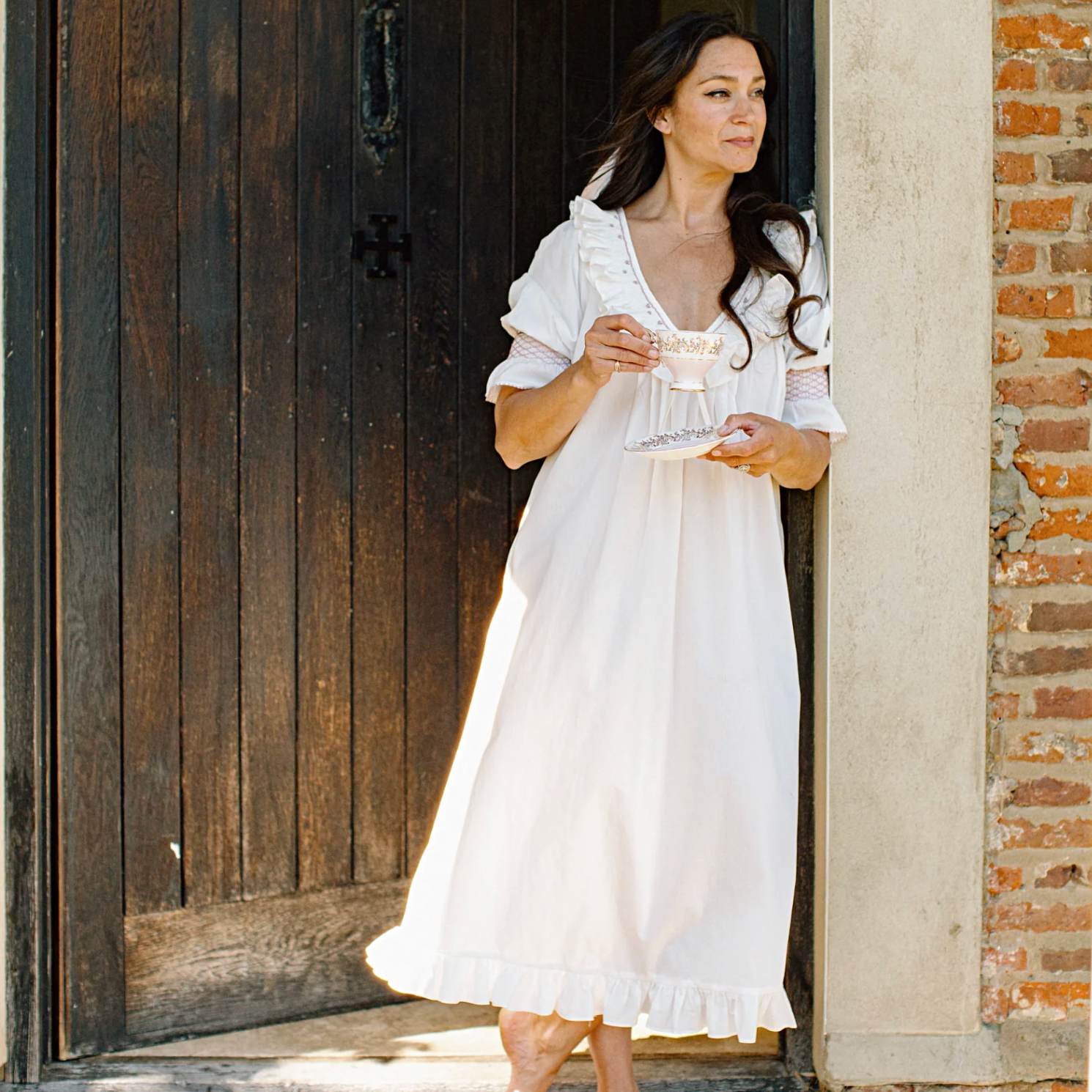 A woman stands barefoot in a doorway wearing a loose white nightgown or dress, holding a small glass cup and saucer. The setting includes a dark wooden door, a brick wall and a light stone column.