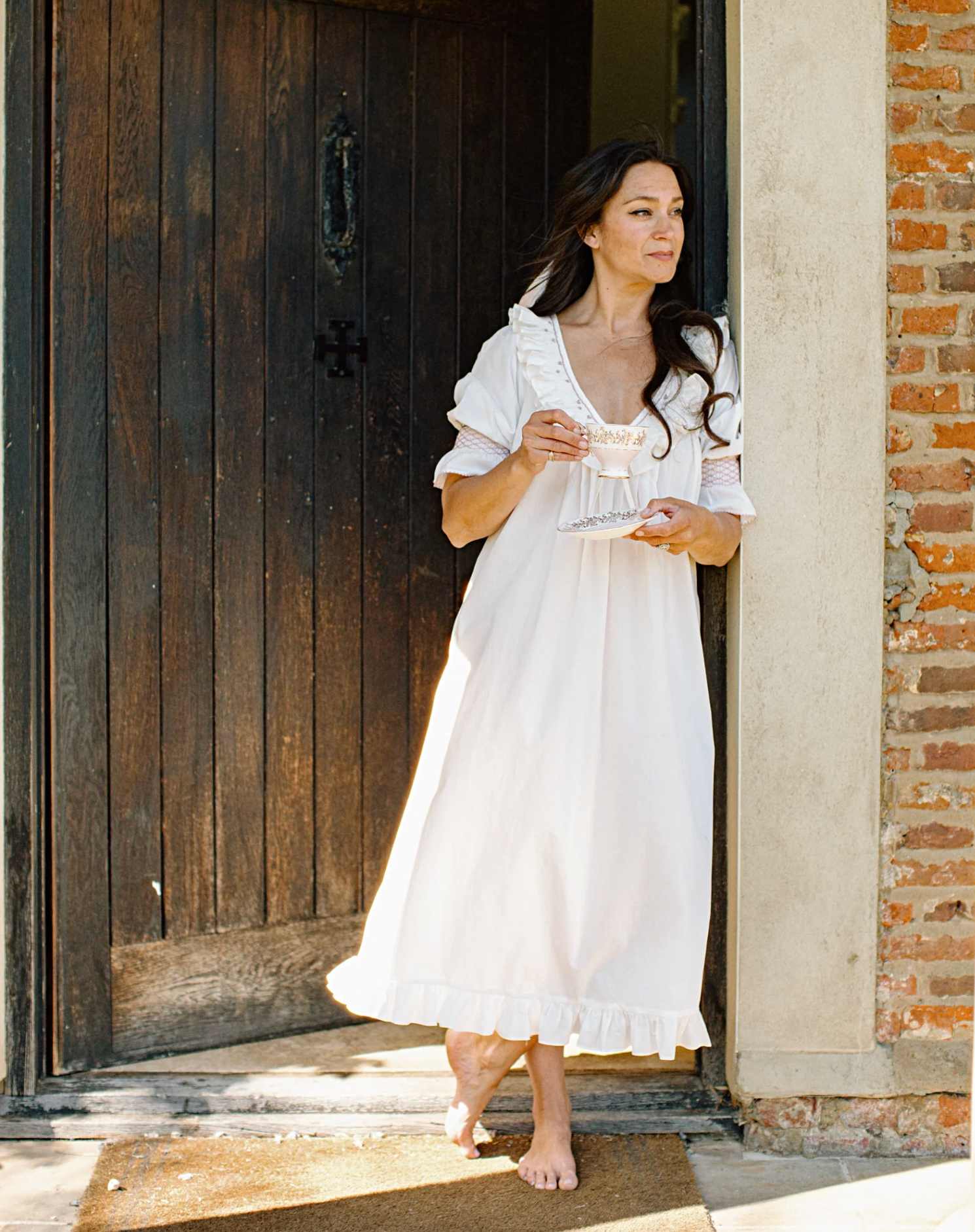 A woman stands barefoot in a doorway wearing a loose white nightgown or dress, holding a small glass cup and saucer. The setting includes a dark wooden door, a brick wall and a light stone column.