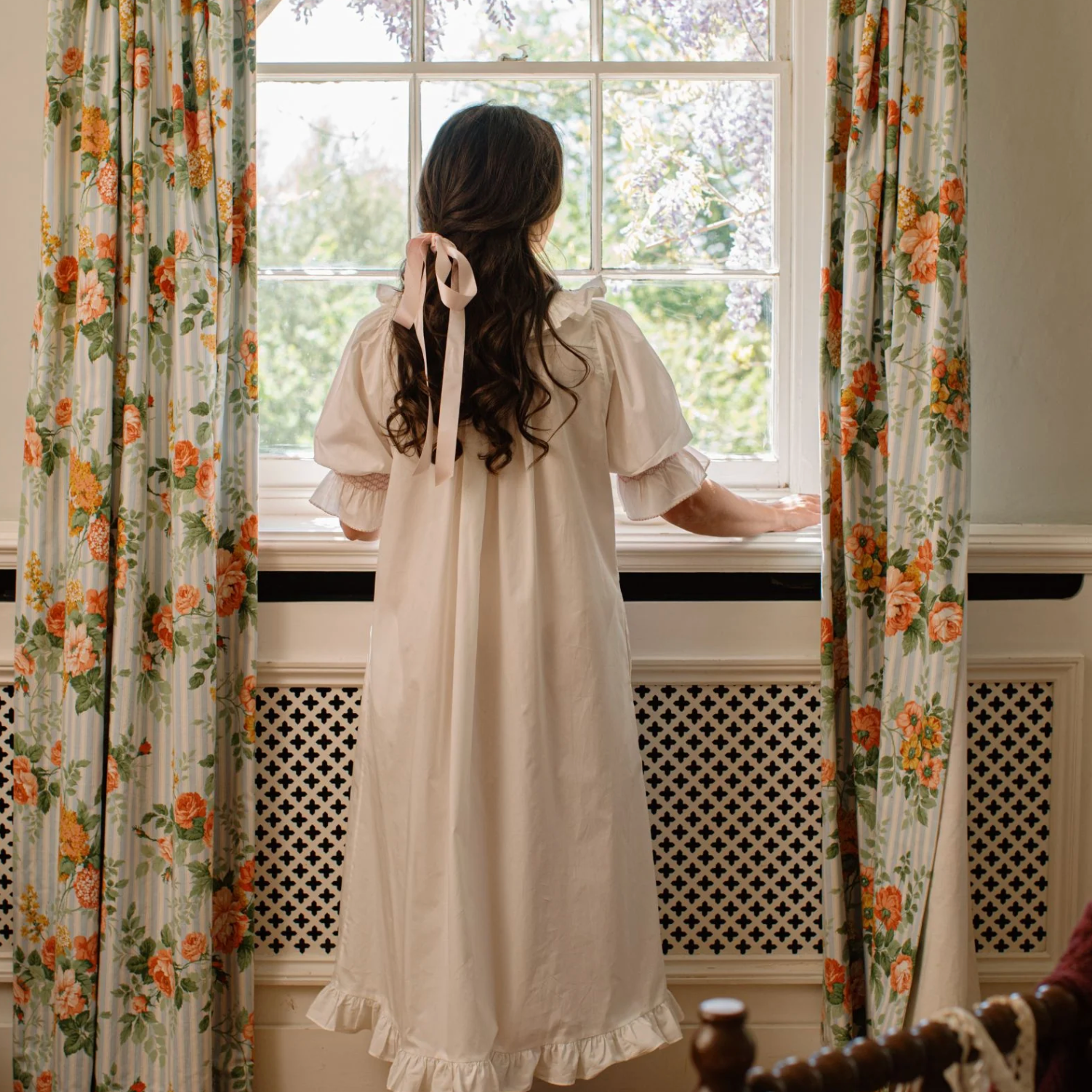A woman viewed from behind wearing a cream-colored, long-sleeved nightgown with a ruffled hem and a matching ribbon bow in her hair, standing by a window with floral curtains.
