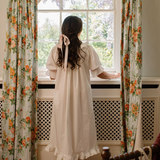 A woman viewed from behind wearing a cream-colored, long-sleeved nightgown with a ruffled hem and a matching ribbon bow in her hair, standing by a window with floral curtains.