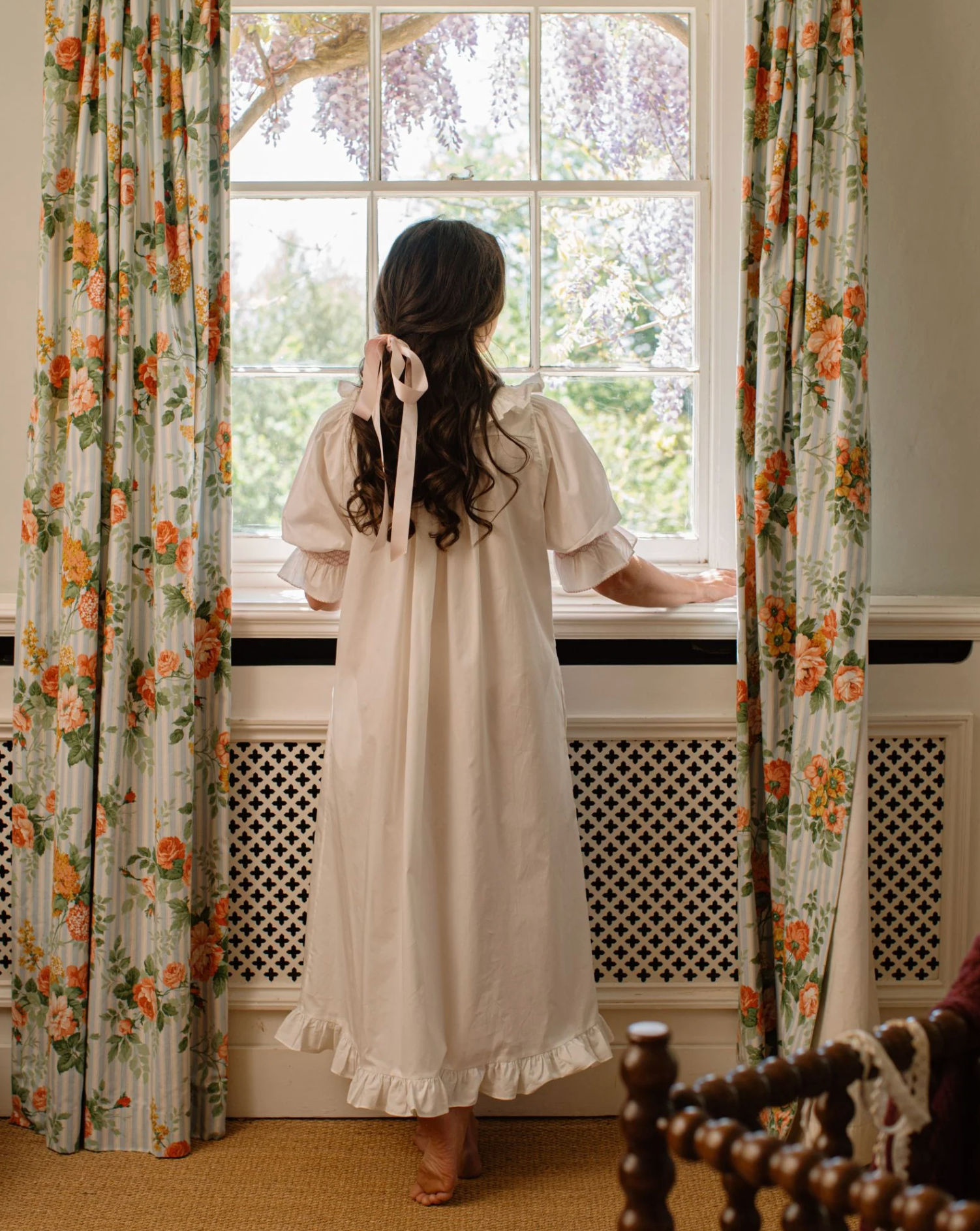 A woman viewed from behind wearing a cream-colored, long-sleeved nightgown with a ruffled hem and a matching ribbon bow in her hair, standing by a window with floral curtains.
