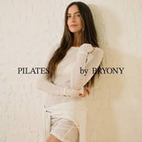 A woman with long dark hair poses in a white long-sleeve top and matching white wrap skirt against a textured white wall. The text 'PILATES by BRYONY' is overlaid on the image.