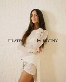 A woman with long dark hair poses in a white long-sleeve top and matching white wrap skirt against a textured white wall. The text 'PILATES by BRYONY' is overlaid on the image.