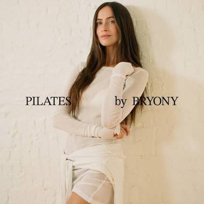 A woman with long dark hair poses in a white long-sleeve top and matching white wrap skirt against a textured white wall. The text 'PILATES by BRYONY' is overlaid on the image.