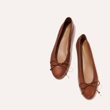 A pair of brown leather ballet flats with small bow details on the toes, photographed from a top-down angle on a light cream background.