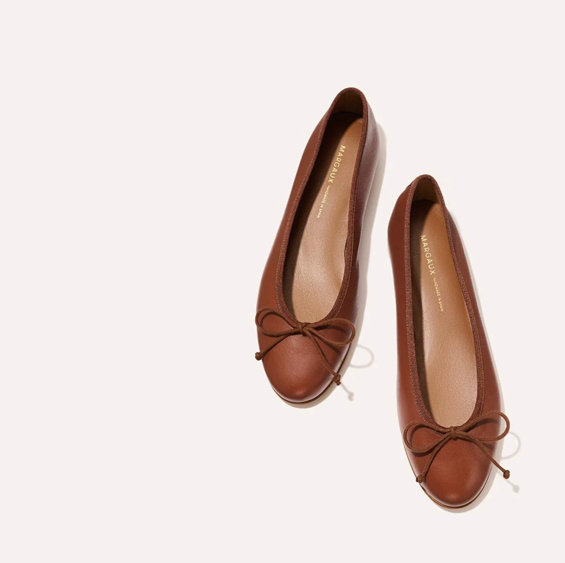 A pair of brown leather ballet flats with small bow details on the toes, photographed from a top-down angle on a light cream background.
