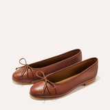 A pair of brown leather ballet flats with small bow details on the toes, photographed on a clean, light beige background.