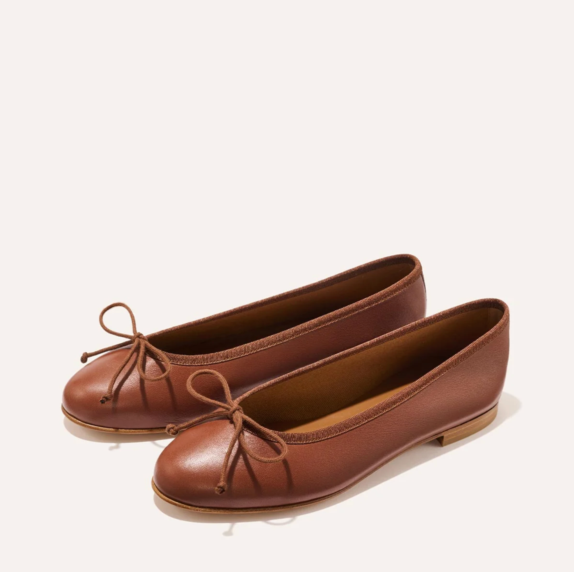 A pair of brown leather ballet flats with small bow details on the toes, photographed on a clean, light beige background.